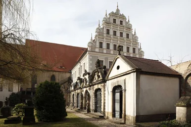Im Vordergrund zu sehen ist ein historisches Grufthaus mit mehreren mit schmiedeeisernen Gittern versehenen Eingängen. Im Hintergrund ragt ein sich über drei Obergeschosse streckender Giebel eines großen spätgotischen Gebäudes auf.