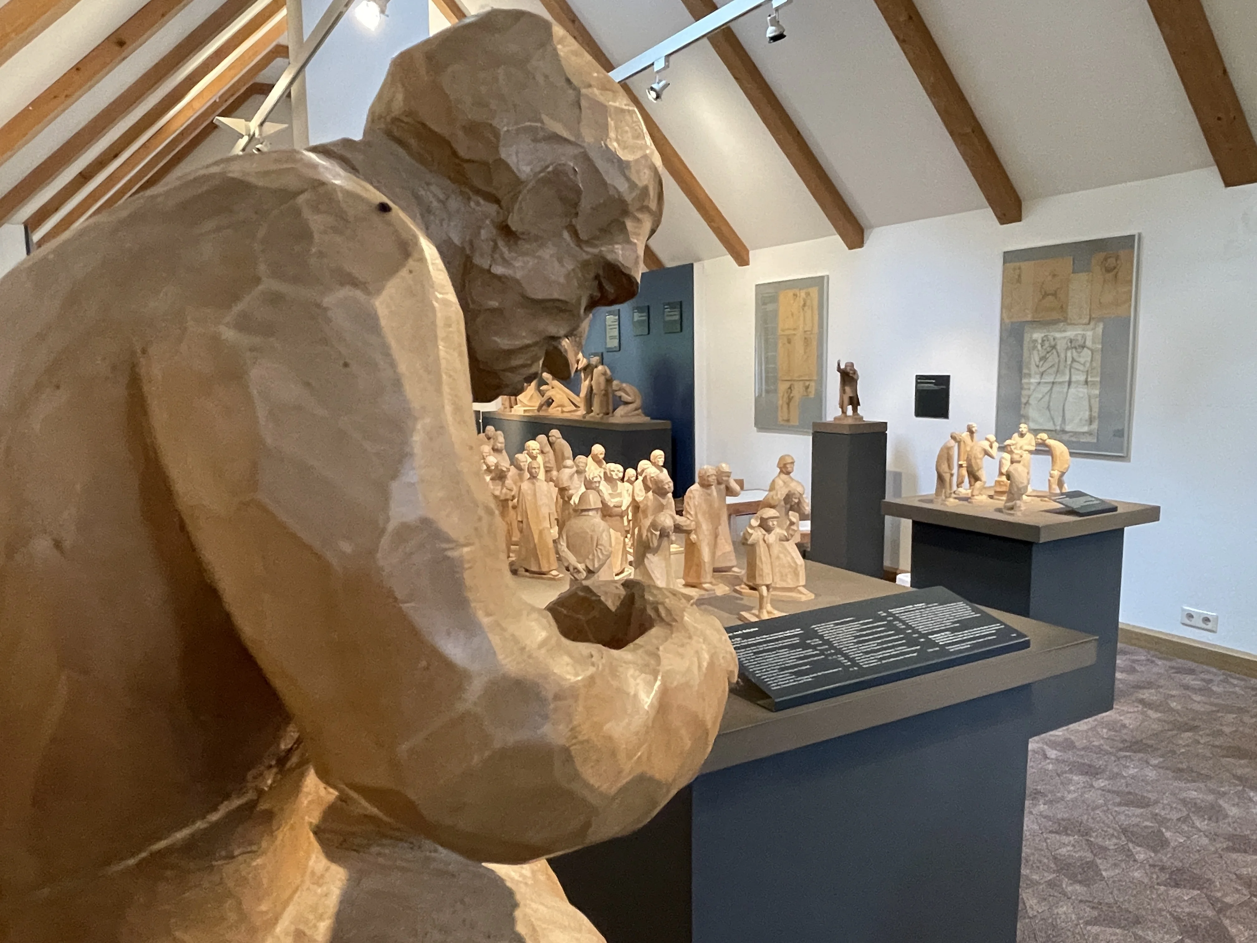 Im Vordergrund steht angeschnitten eine große Holzfigur, dahinter weitere geschnitzte Werke in einem hellen Ausstellungsraum.