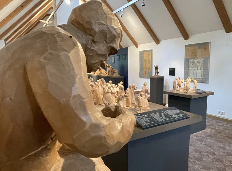 Im Vordergrund steht angeschnitten eine große Holzfigur, dahinter weitere geschnitzte Werke in einem hellen Ausstellungsraum.