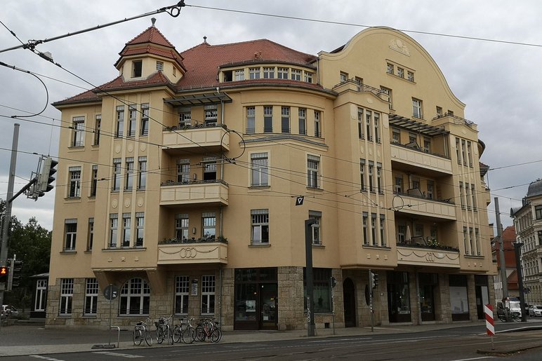 Ein großes, vierstöckiges Eckhaus im Stil der Gründerzeit mit gelber Fassade, Balkonen und einem Türmchen