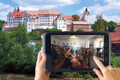 Mit dem HistoPad Schloss Colditz entdecken Im Vordergrund wird von zwei Händen ein Tablet-Guide gehalten, im Hintergrund sind ein großes Schloss und ein darunter verlaufender Fluss zu sehen.