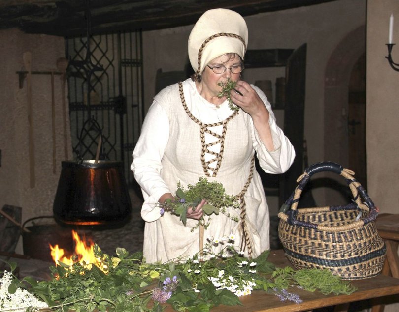 Eine im historischen Kostüm einer Köchin gekleidete Frau riecht an einer Handvoll Kräuter. Vor ihr auf einem Tisch ausgebreitet liegen weitere Kräuter und Blumen sowie ein Korb. An der alten Herdstelle hinter ihr lodert ein Feuer.