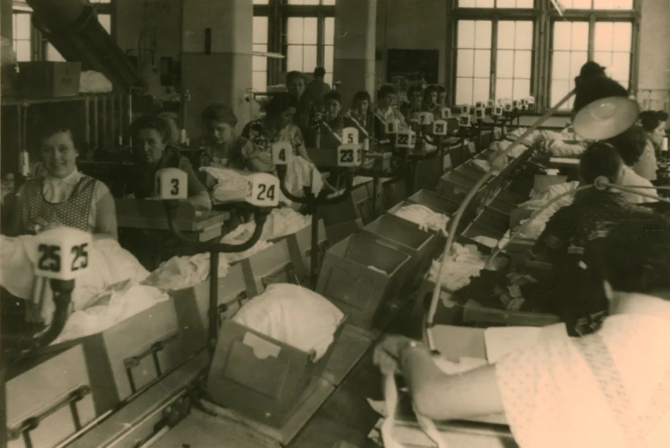 Die historische, sepia-farbene Fotografie zeigt Näherinnen in einen großen Näh-Saal.