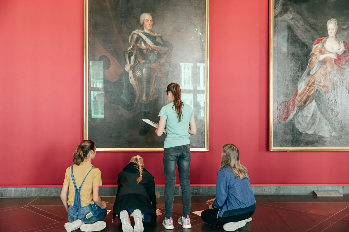 Das Foto zeigt vier Schülerinnen von hinten, die, mit Stift und Papier ausgestattet, das große Porträt eines sächsischen Herrschers aus der Barockzeit betrachten. Rechts von diesem Gemälde hängt ein großes Porträt einer weiblichen Person aus dieser Zeit.
