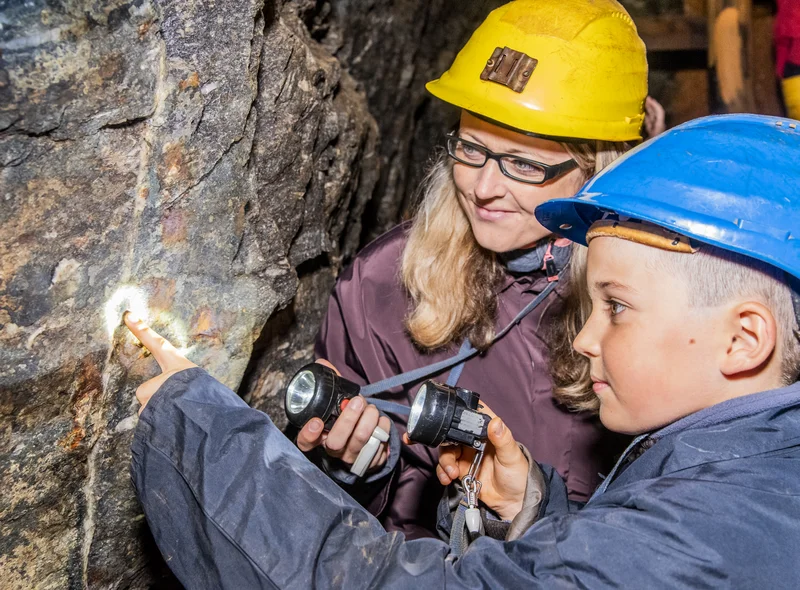 In einem Stollen stehen eine Frau und ein Kind in Schutzkleidung (Overall und Schutzhelm) vor einer Steinwand. Beide halten eine kleine Taschenlampe in der Hand und richten diese auf einen hellen, vertikalen Streifen im dunklen Gestein der Wand. Das Kind zeigt mit der freien Hand auf einen Punkt in diesem Streifen, den beide anleuchten.