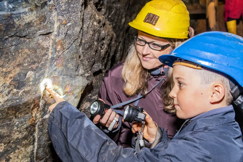 In einem Stollen stehen eine Frau und ein Kind in Schutzkleidung (Overall und Schutzhelm) vor einer Steinwand. Beide halten eine kleine Taschenlampe in der Hand und richten diese auf einen hellen, vertikalen Streifen im dunklen Gestein der Wand. Das Kind zeigt mit der freien Hand auf einen Punkt in diesem Streifen, den beide anleuchten.