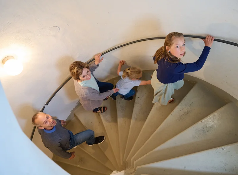 Eine Familie steigt die Stufen einer alten Wendeltreppe aus Stein an einem Geländer nach oben.