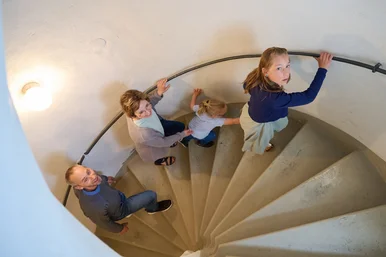 Ferienprogramm „Steinalt“ im Schloss Museum Glauchau Eine Familie steigt die Stufen einer alten Wendeltreppe aus Stein an einem Geländer nach oben.