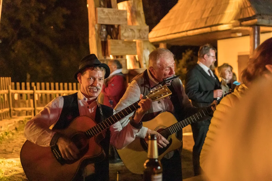 Am Rande einer Menschengruppe spielen zwei Musiker Gitarre und Mundharmonika. Sie stehen im Freien, es ist dunkel.