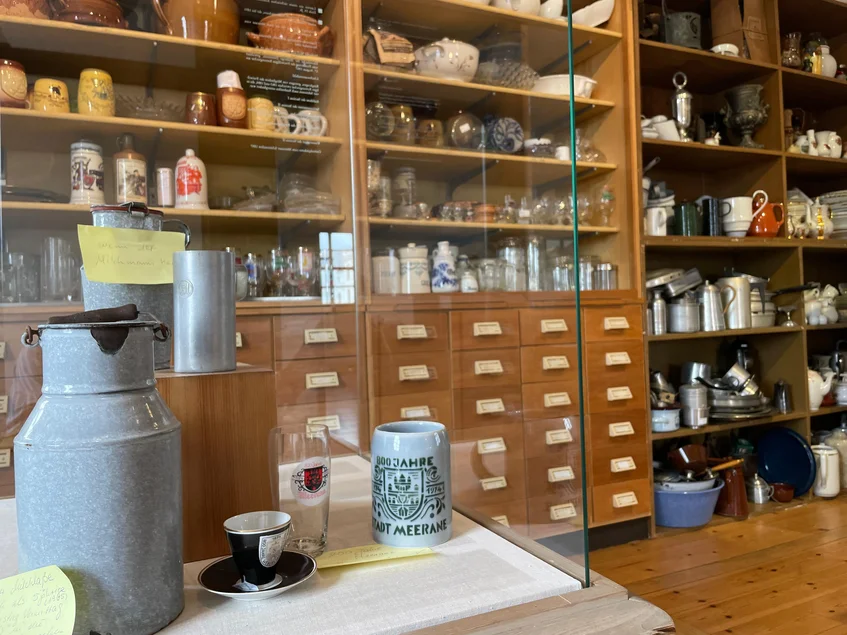 Einige bunt zusammengewürfelte Objekte stehen in einer Vitrine, dahinter ein großes, chaotisch gefülltes Regalsystem.