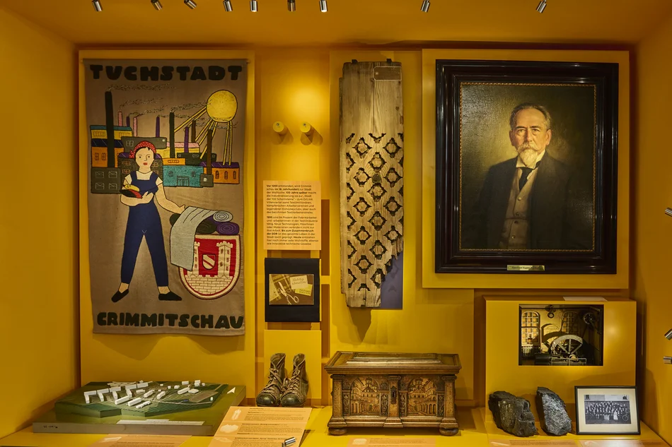 Vitrinenansicht In der Vitrine sind ein gewebter Wandbehang mit einer Industriestadtansicht und den Worten "Tuchstadt Crimmitschau", das Porträt eines älteren Mannes, eine kleine Truhe, ein paar alter Lederschuhe und weitere kleinere Exponate ausgestellt.