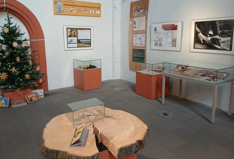 Ausstellungsraum mit einer Baumstammscheibe, mehreren Wandbildern und Vitrinen sowie einer mit Sternen geschmückten Tanne