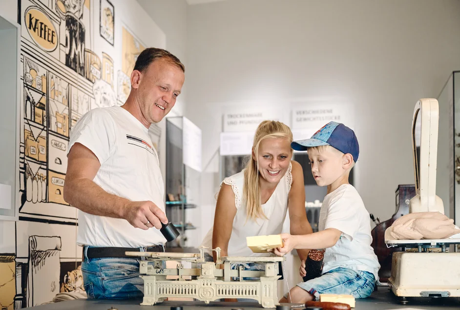 Ein Mann, eine Frau und ein kleiner Junge probieren in einem als Krämerladen gestalteten Mitmachbereich der Ausstellung das Wiegen mit einer historischen historischen Balkenwaage aus.