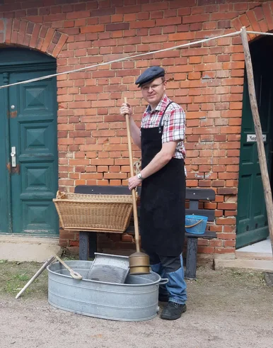 Ein Mann mit brauner Schürze steht neben einer Zinkwanne mit Waschbrett und anderen historischen Wasch-Utensilien, hinter ihm auf einer Bank ein geflochtener Wäschekorb.