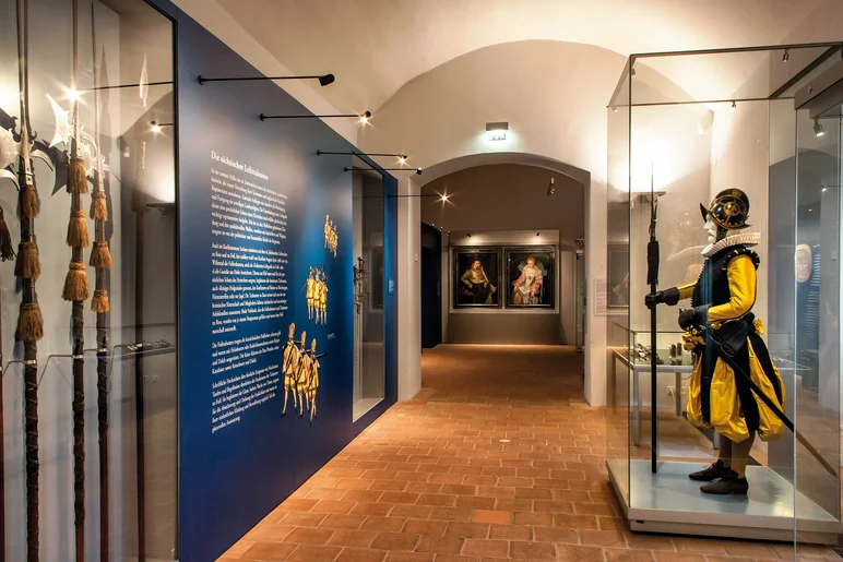 In dem länglichen Ausstellungsraum befinden sich linkerhand eine Vitrine mit historischen Stichwaffen sowie ein Wandtext und rechterhand in einer Vitrine die lebensgroße Figur eines Mannes in Prachtuniform. An der Stirnseite des Raumes hängen zwei historische Gemälde.