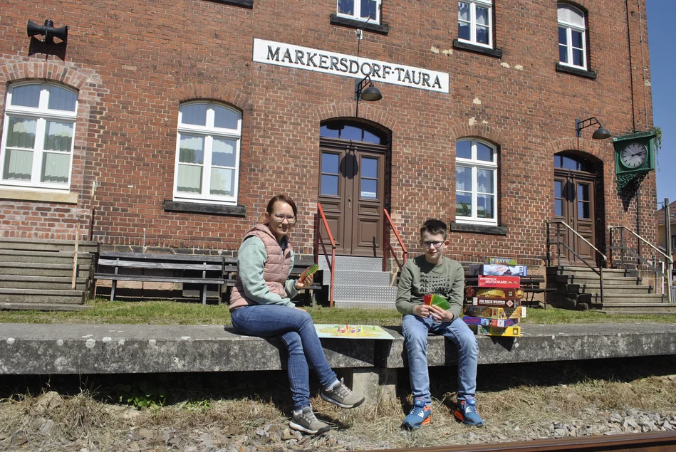 Ein Mädchen und ein Junge sitzen auf der Bahnsteigkante des Museumsbahnhofs Markersdorf-Taura und spielen ein Brettspiel.