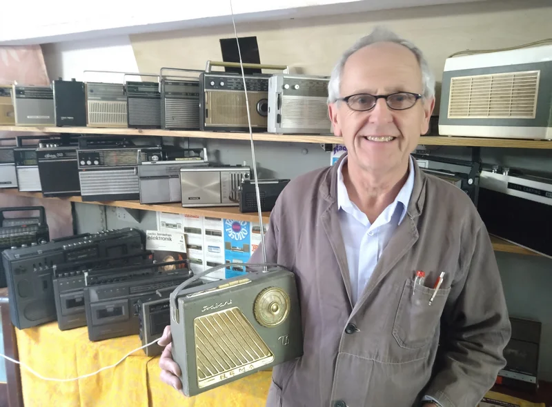 In einem mit historischen Radios bestückten Ausstellungsraum zeigt ein Mann in grauem Arbeitskittel ein altes Kofferradio.