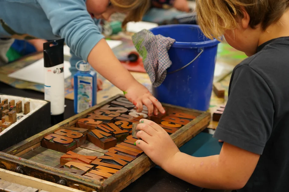 Zwei sich gegenübersitzende Kinder greifen in einen zwischen ihnen auf einem Arbeitstisch liegenden Setzkasten mit großen hölzernen Druckbuchstaben. Daneben steht ein blauer Eimer mit Lappen.