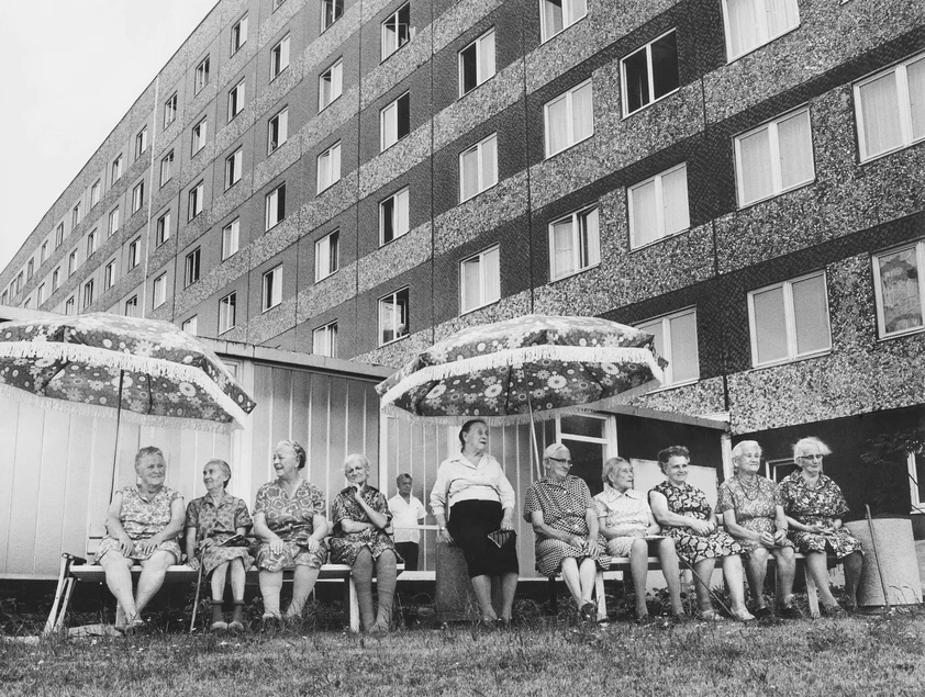 Dresden-Mockritz, Altersheim Olga Körner, um 1974 Schwarzweißfotografie eines Plattenbaus, vor dem unter zwei Sonnenschirmen mehrere ältere Frauen in Kittelschürzen sitzen