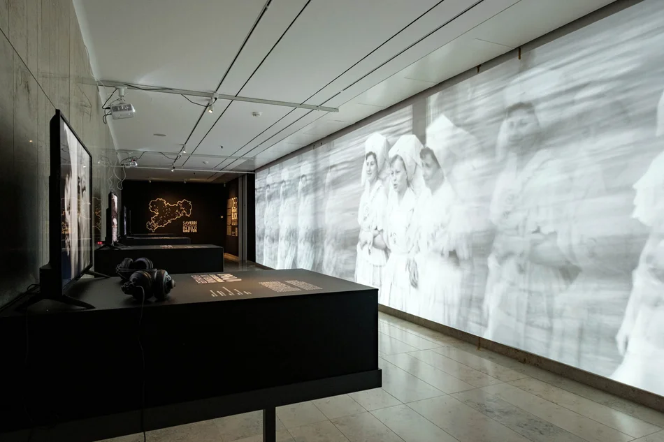 Auf dem Foto ist der Außenbereich des Buchmuseums abgebildet, sozusagen die Galerie, in deren Innerem sich wie ein Kubus die Schatzkammer befindet. Der Raum ist auf dieser Aufnahme dunkel mit Lichtakzenten. Rechts an der Wand ist eine große Filmprojektion zu erkennen, links befinden sich vier Flachvitrinen, auf denen jeweils Bildschirme mit Filmen aufgestellt sind. An der hinteren Wand ist in Leuchtoptik eine Karte von Sachsen zu erkennen.