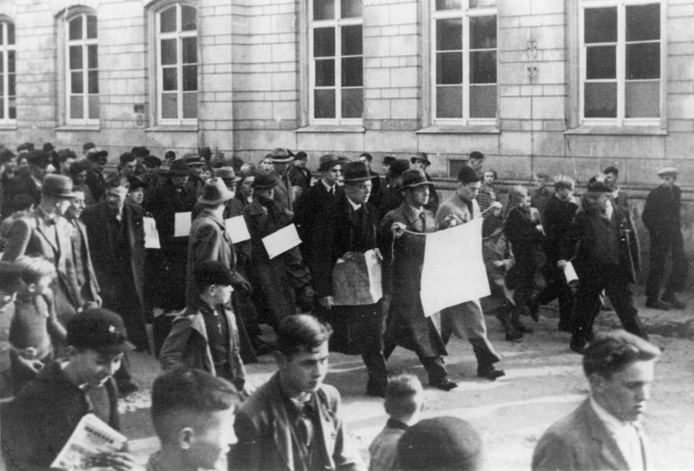 Diese historische Schwarzweiß-Aufnahme von 1938 zeigt, wie jüdische Bürgerinnen und Bürger in Bautzen gezwungen werden, mit von Nazis beschriebenen Plakaten durch die Straßen zu ziehen. Begleitet werden sie dabei seitlich von nicht-jüdischen Jugendlichen und Erwachsenen.