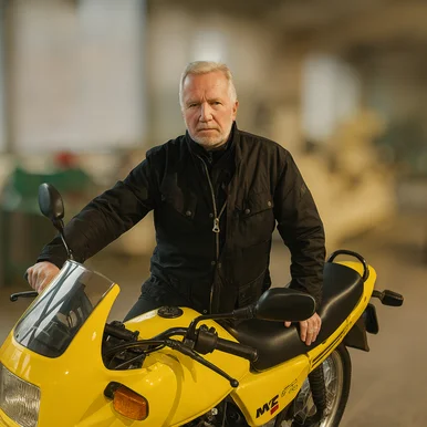 Ein sportlich wirkender älterer Herr mit grauen Haaren und Bart steht an einem gelben Motorrad mit schwarzem Sitz.