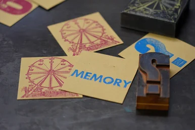 Auf einer Arbeitsfläche liegen vier quadratische Karten (zwei davon sind mit einem identischen Riesenrad-Motiv bedruckt), daneben liegen Hochdruck-Klischees aus Holz.