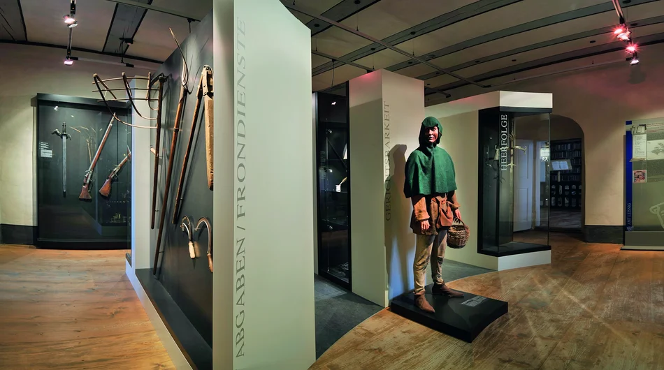 Ausstellungsteil Amtsgeschichte Vorderschloss Blick in einen Ausstellungsraum mit Vitrinen und Stellwänden; zentral ist eine lebensgroße, in einfache mittelalterliche Kleidung gekleidete Figur mit einem Körbchen in der Hand zu sehen. In der Vitrine ganz links befinden sich historische Schusswaffen, an der Stellwand daneben sind alte landwirtschaftliche Geräte angebracht.