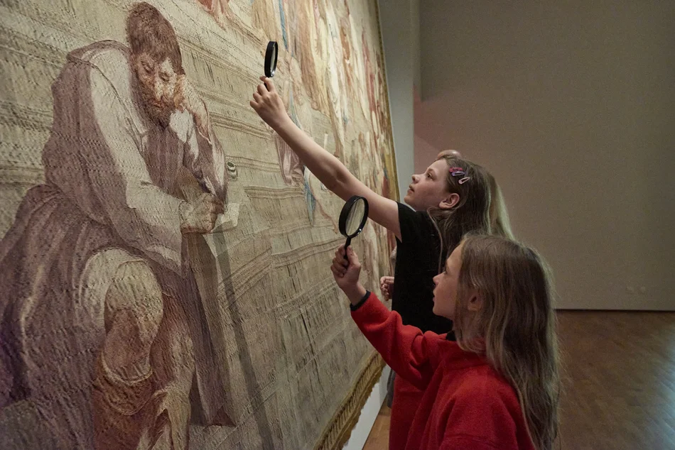 Zwei Kinder betrachten mit einer Lupe einen großen Wandteppich in einer Ausstellung.