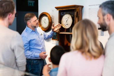 Ein Uhrmachermeister erklärt einer Besuchergruppe das Zifferblatt einer historischen Standuhr.