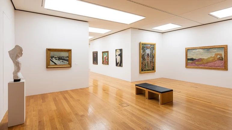 Ausstellungsraum mit weißen Wänden, an denen diverse Gemälde hängen, linkerhand eine abstrakte Büste auf einem Podest, mittig eine Bank zum Verweilen