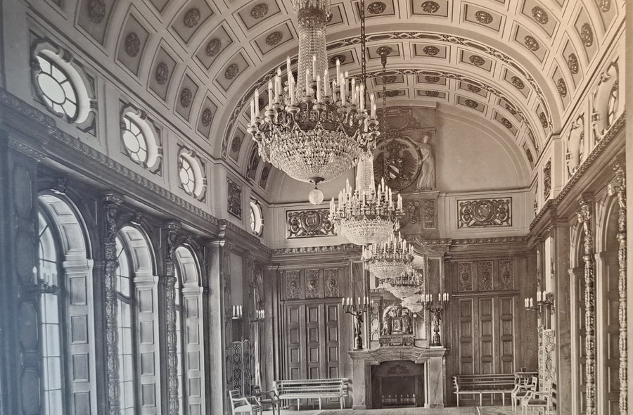 Historisches Foto in Sepia-Tönen, das einen prunkvollen Saal mit hoher Decke und prachtvollen Kronleuchtern zeigt