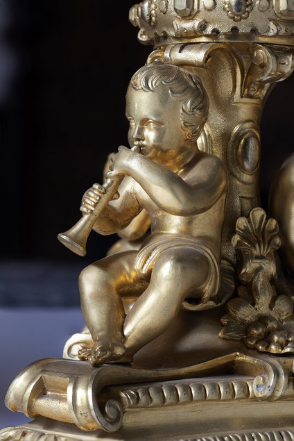 Eine goldverzierte kleine Kindergestalt sitzt, angelehnt an ein reich verzierte Säule, und spielt ein Blasinstrument.