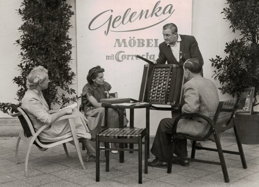 Historisches Schwarzweißfoto, auf dem vor einer Wand mit dem Schriftzug "Gelenka – Möbel mit Correcta" ein Mann in dunklem Anzug drei auf Armlehnstühlen sitzenden Personen ein weiteres Sitzmöbel zeigt