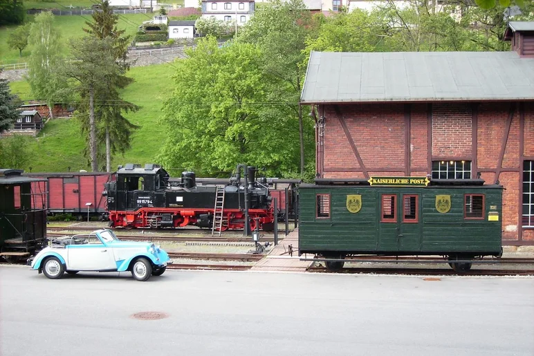 Neben einem kleinen alten Ziegelsteingebäude stehen eine Dampflok und historische Waggons, davor ein Oldtimer-Cabriolet.
