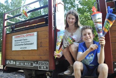 Zuckertüten-Express im Chemnitztal Ein Mädchen und ein Junge sitzen im Einstiegsbereich eines offenen Eisenbahnwaggons und halten je zwei kleine Zuckertüten in den Händen.