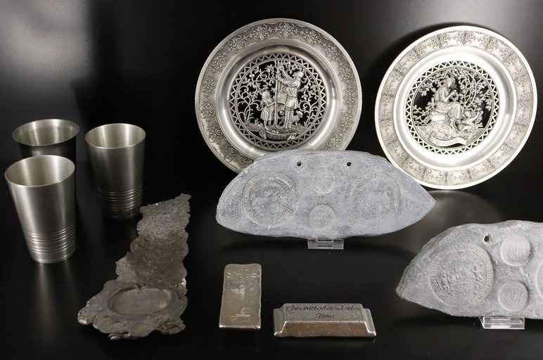 Inhalt einer Museumsvitrine mit zwei Zinntellern, zwei Zinnbarren, drei Zinnbechern, einem langen Stück geschmolzenen Zinn und zwei Gussformen für Zinnmünzen.