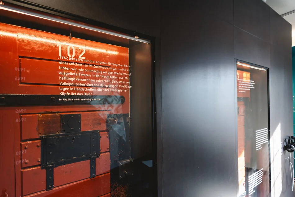 Nahaufnahme einer Wandvitrine, in der sich eine historische Gefängniszellentür mit der Nummer 102 befindet. Auf das Vitrinenglas ist das Zitat eines politischen Häftlings der 1960er Jahre gedruckt.