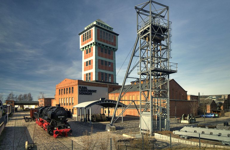 Foto eines Klinker-Ensembles mit Förderturm und sich anschließenden niedrigeren Industriebauten, daneben steht ein weiterer, aus Stahl konstruierter Turm.