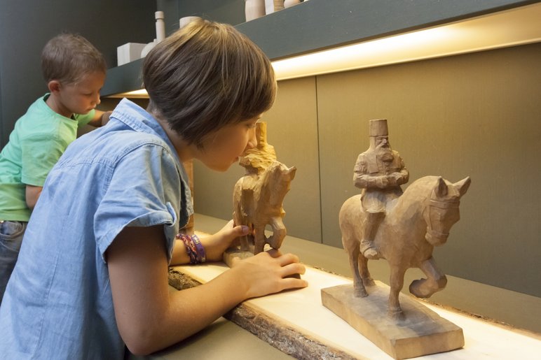 Das Foto zeigt zwei Kinder, die handgeschnitzte Holzobjekte – jeweils ein auf einem Pferd reitender Bergmann – aus der Nähe betrachten.