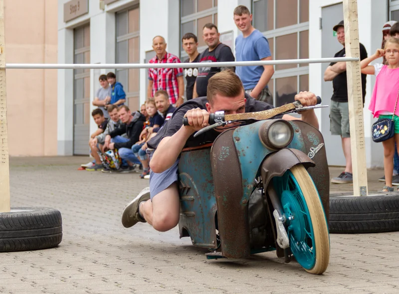 Ein Mann auf einem historischen Simson-Motorroller fährt unter einer niedrig gehängten Stange hindurch.