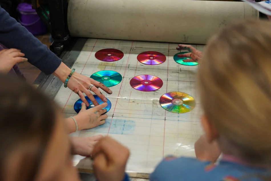 Mehrere zu Tiefdruckformen umgewandelte Compact-Disk-Tonträger liegen mit der Unterseite nach oben auf einem zu bedruckenden Material, dahinter ist der Zylinder einer Druckpresse zu sehen.
