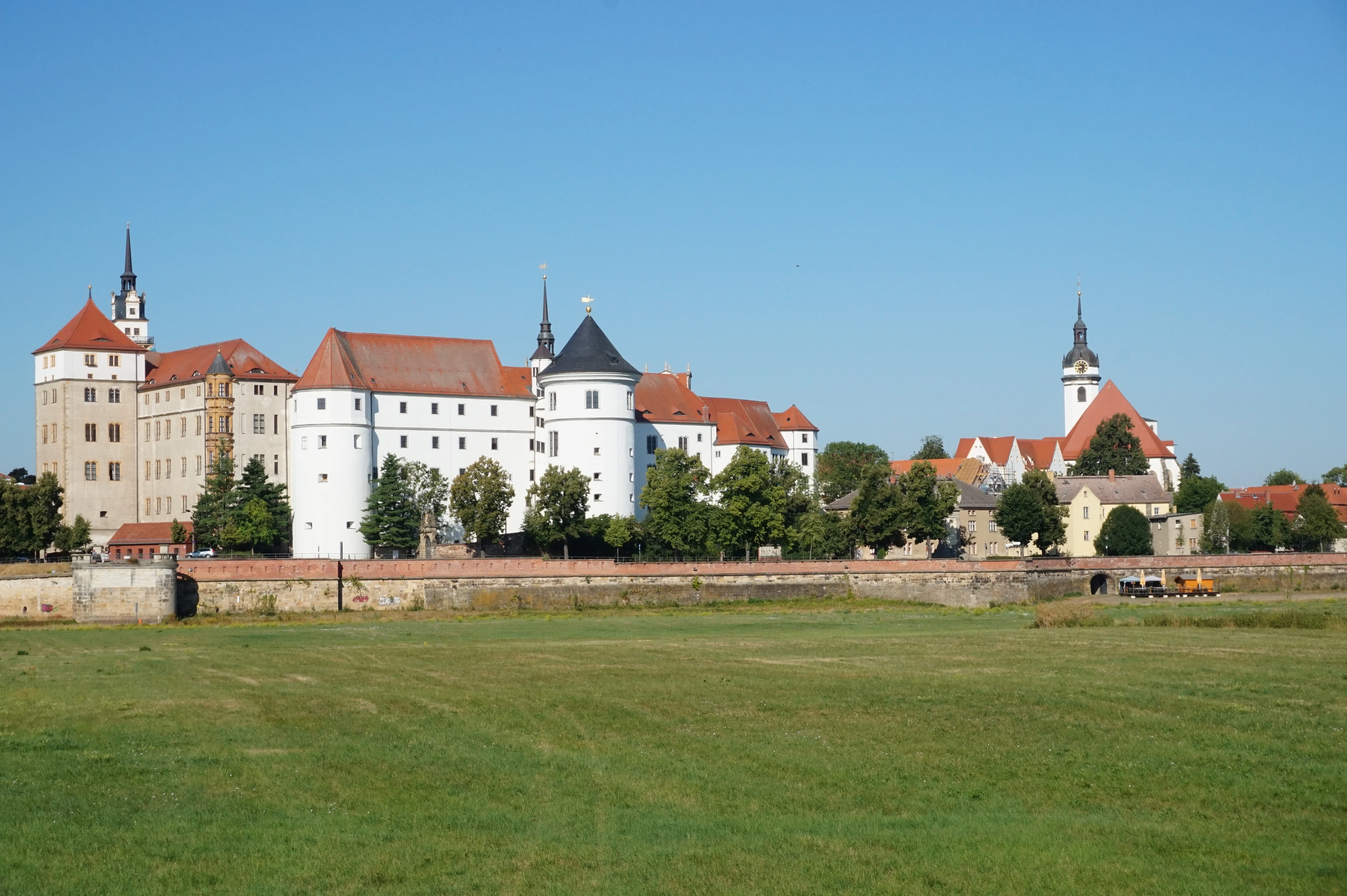 Blick über die Elbwiesen auf Schloss Hartenfels Sommerliche Außenansicht eines großen Schlosskomplexes mit weißer Fassade und mehreren Türmen, im Vordergrund eine flache Grasfläche