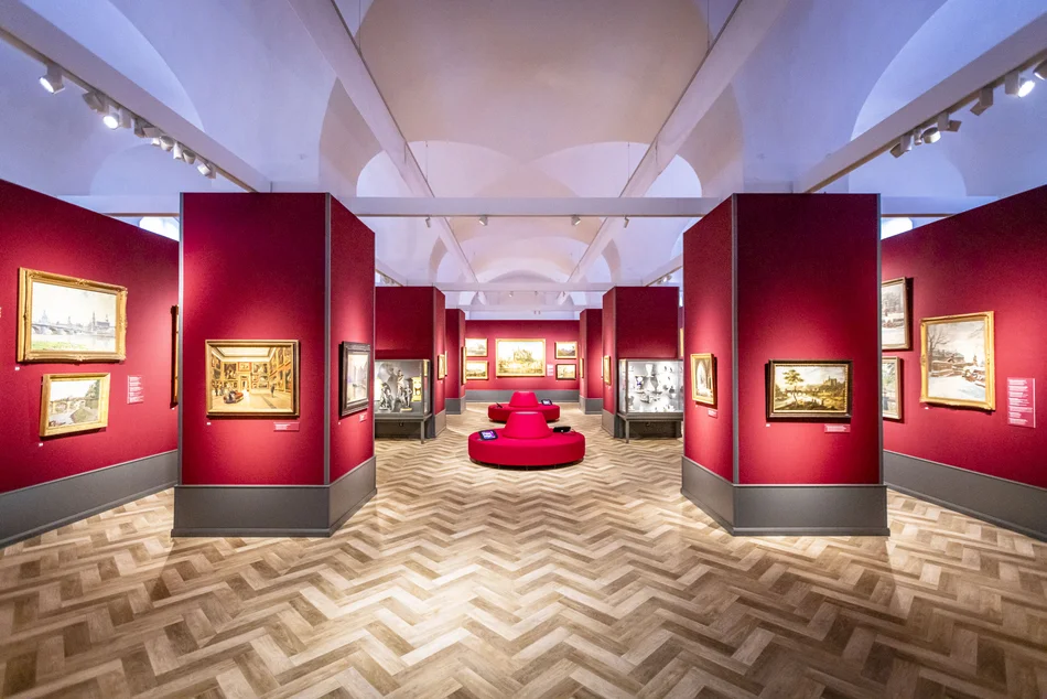 Blick in einen edel wirkenden Saal mit weinroten Ausstellungswänden, Gemälden und Vitrinen mit Skulpturen und Porzellan