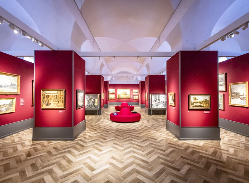 Blick in einen edel wirkenden Saal mit weinroten Ausstellungswänden, Gemälden und Vitrinen mit Skulpturen und Porzellan
