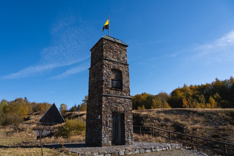 Ein gemauerter Turm mit Eisentor vor Haldengelände und blauem Himmel. Auf dem Turm ist ein Wimpel in gelb-schwarz angebracht.