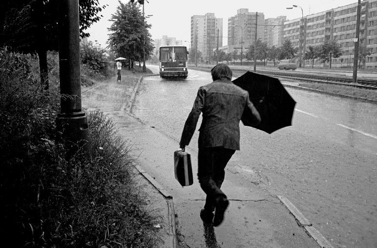 Schwarzweißfotografie eines Mannes mit Schirm, der in einem Neubaugebiet durch den Regen auf eine Bushaltestelle zuläuft