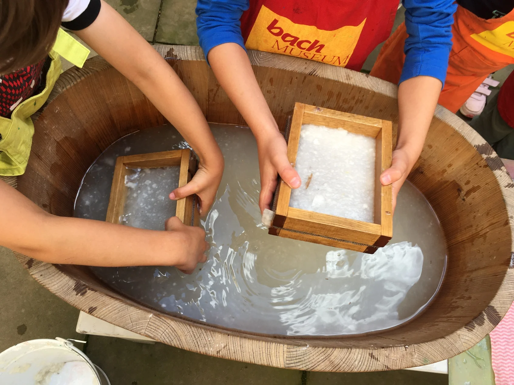 Das Foto zeigt die Arme und Hände zweier Kinder, die über einem Bottich mit Pulpe einen Rahmen halten und Papier schöpfen.