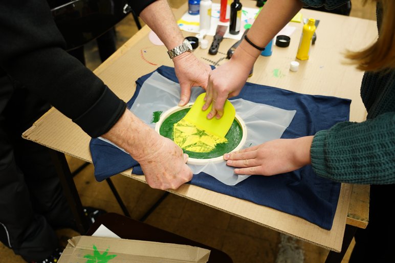 Zwei Hände fixieren einen runden Stickrahmen auf einer Arbeitsfläche, zwei andere Hände drücken mit einer Art Spatel grüne Farbe durch das in Gewebe auf ein blaues Textil.
