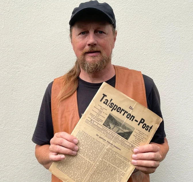 Der Referent Eckhart Böhm mit der Zeitschrift „Die Talsperren-Post“ Eckhart Böhm, ein Mann mittleren Alters mit Kappe und Zopf, hält die historische Zeitschrift Die Talsperren-Post in der Hand
